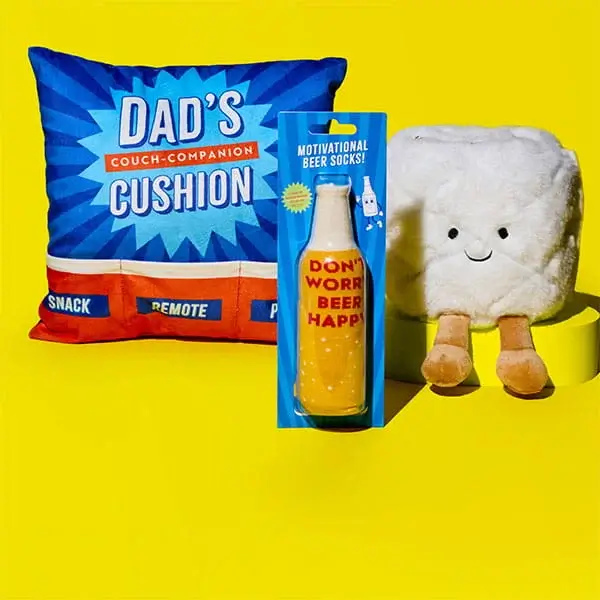 Fathers Day gift display on a yellow background featuring a Dad couch companion cushion, novelty beer bottle socks, and a plush character toy